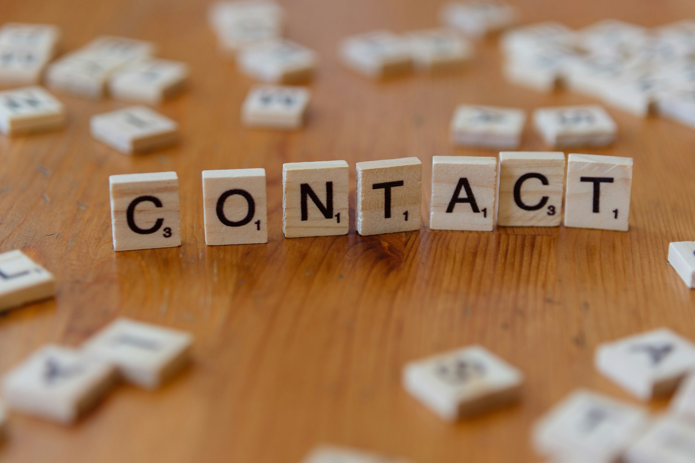 Holzbuchstaben auf einem Holztisch, aufgestellt zum englischen Wort „CONTACT“, im Hintergrund unscharf weitere lose Buchstabensteine