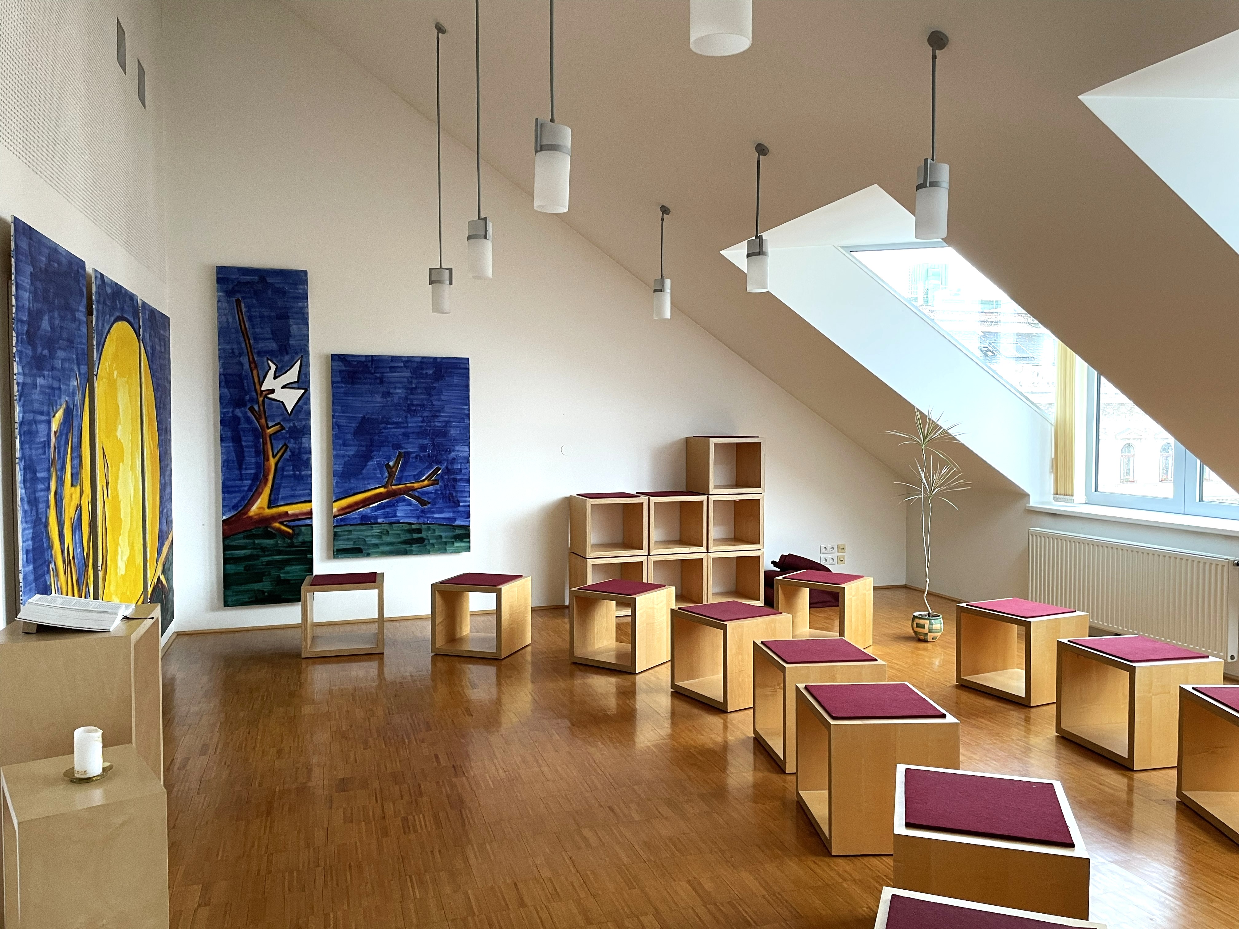 Auf dem Bild sieht man den Andachtsraum. Man sieht auf der linken Seite das Wandbild, welches sich auf der danebenligenden Wand fortsetzt. Gegenüber von dem Wandbild befinden sich in zwei Reihen aufgestellt Hocker. Von der Decke hängen mehrere Lampen