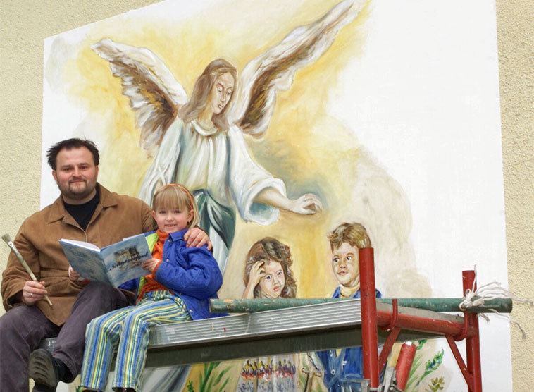 Der Künstler Franz Trink mit seiner Tochter hat 2001 ein SchutzEngel-Bild an die Fassade des Pfarrkindergartens gemalt. Inspiriert zu dem Motiv hat ihn eine Abbildung aus Günther Nennings Buch über die Engel.