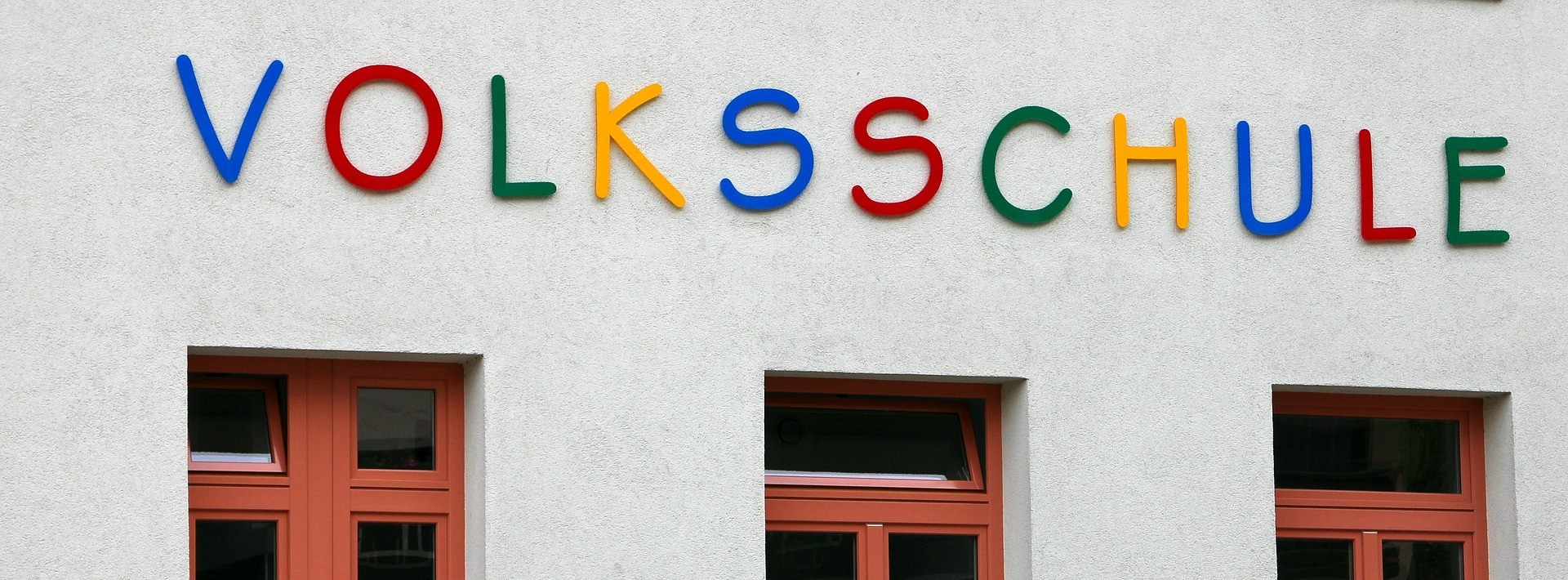 Auf dem Bild sieht man einen Ausschnitt von einer Volksschule. Man sieht den oberen Teil von Fenster und prominent in der Mitte den bunten Schriftzug 'Volksschule'.
