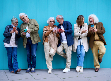 Group of six elderly Caucasian happy people leaning on blue wall using mobile phone. Cheerful mature grey hair couples friends together having fun enjoying looking smiling cellular telephone outdoors