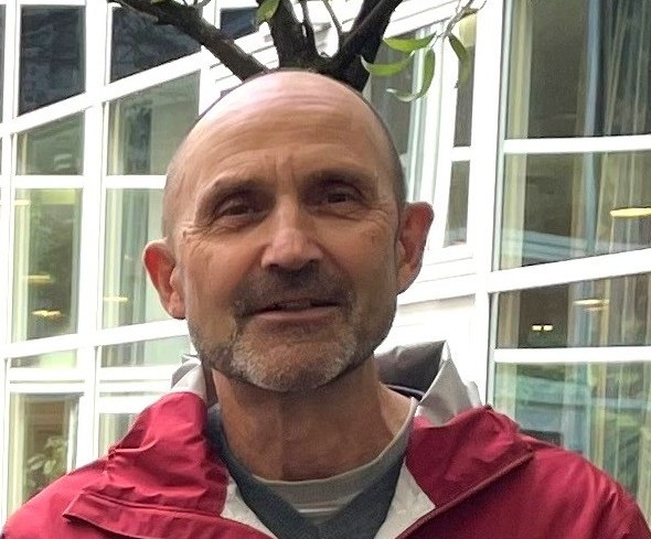 Auf dem Bild sieht man einen Mann mit kurzen Haaren und einer roten Jacke. Er steht vor einer Fensterfront.