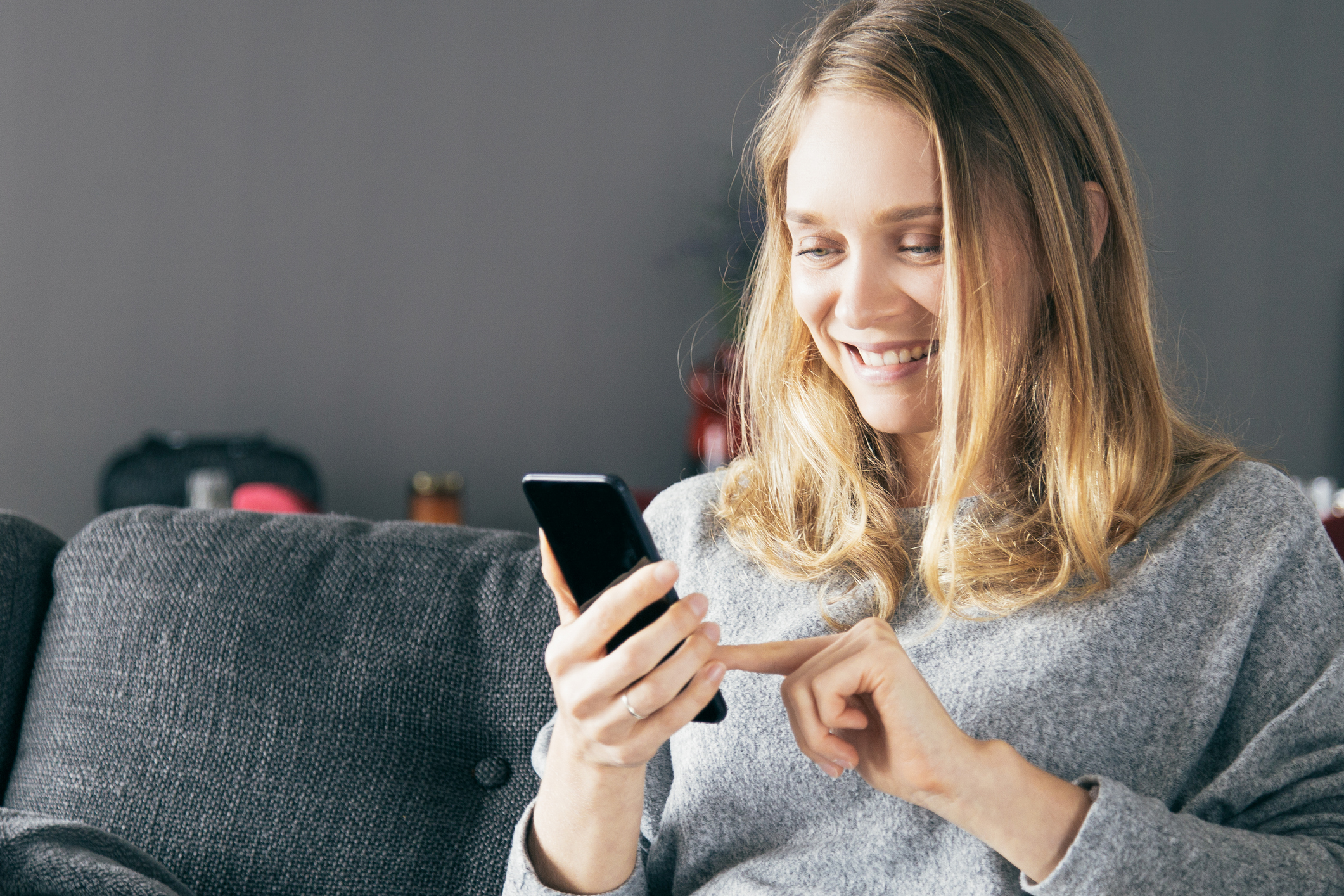 Positive Caucasian girl happy about good news. Beautiful young woman resting on couch at home, using phone and smiling at screen. Online service concept