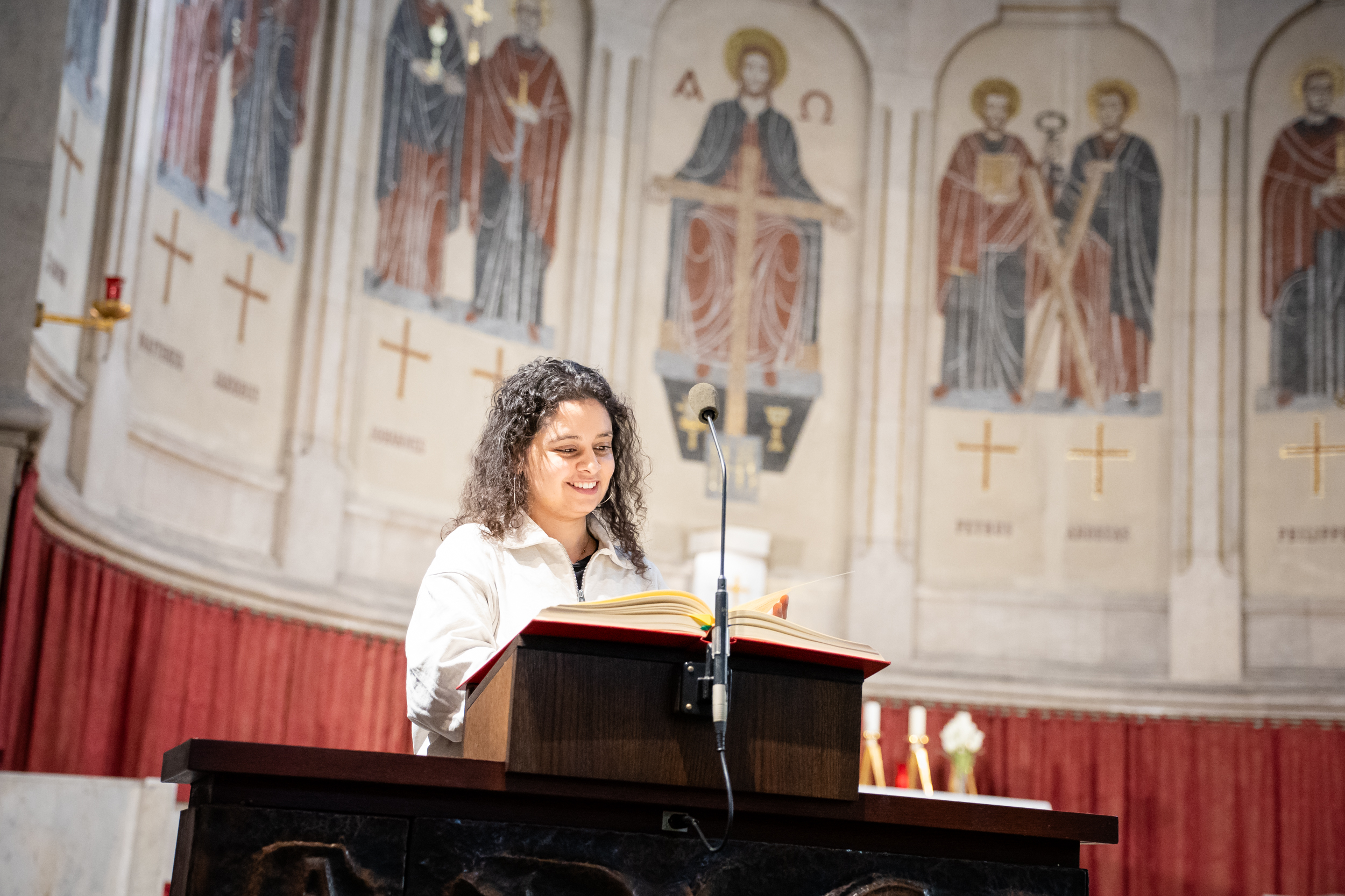 Auf dem Bild sieht man eine junge Pastoralassistentin, die in einer Kirche am Redepult steht und etwas vorliest. Im Hintergund sind an die Kirchenwand verschiedene Heilige aufgezeichnet.