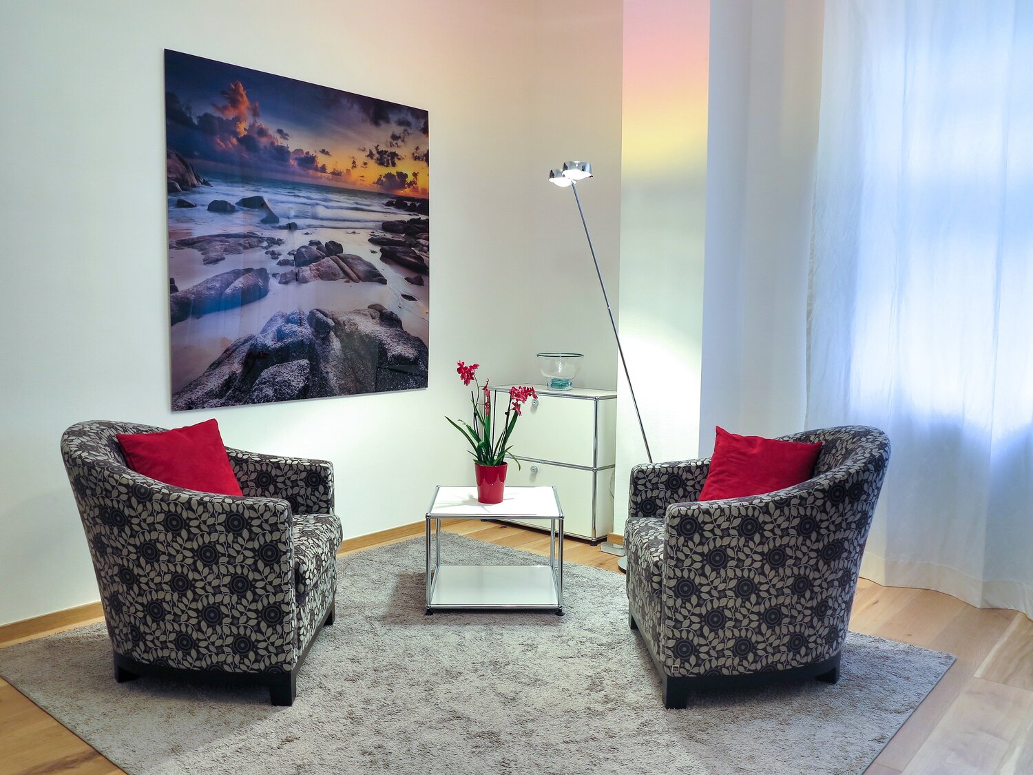 Zwei gemusterte Sessel mit roten Kissen stehen auf einem Teppich an einem kleinen Tisch mit roter Orchidee, daneben eine Stehlampe und ein großes Wandbild