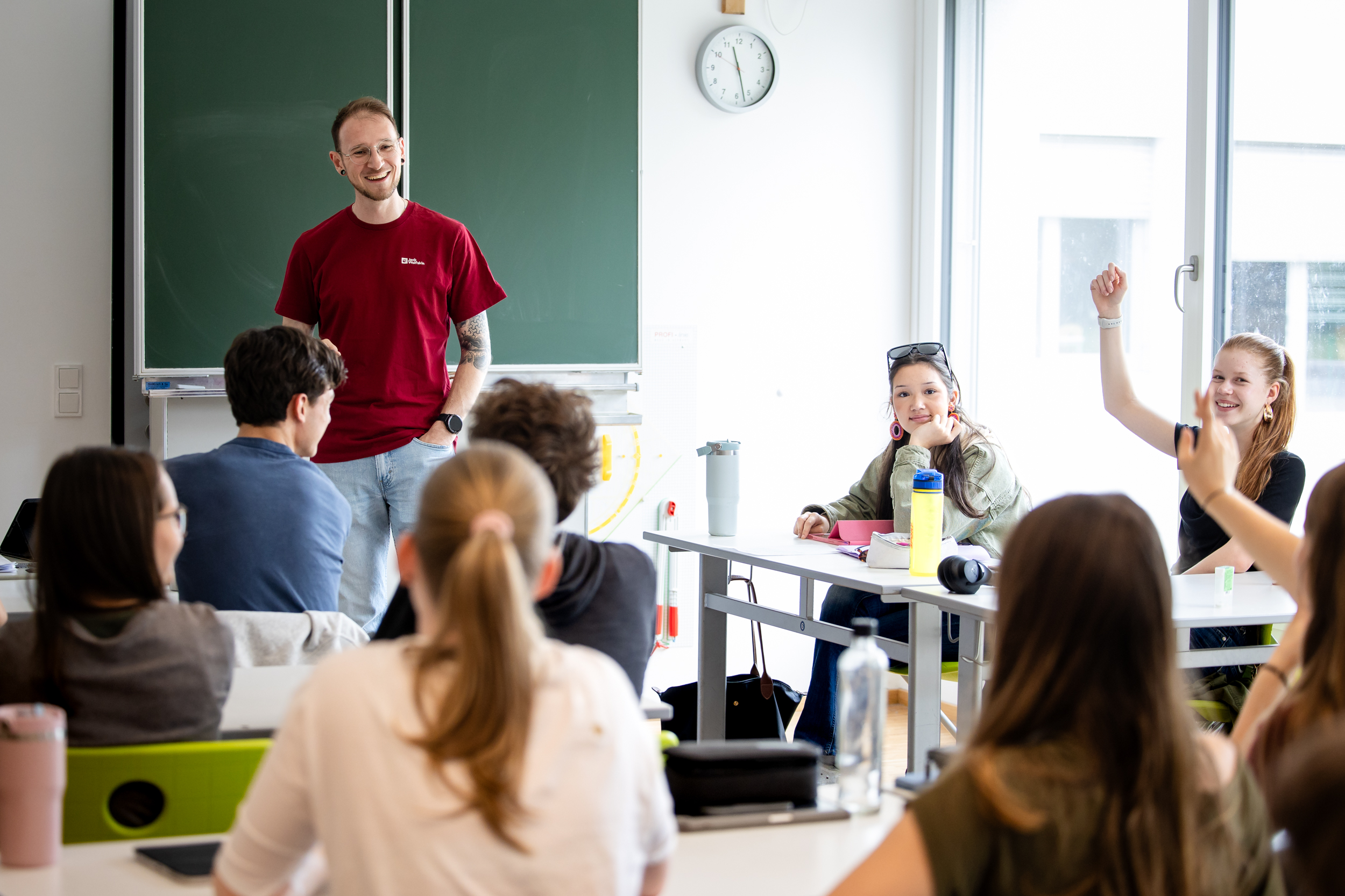 Auf dem Bild sieht man einen Religionslehrer mit einem roten Pullover und einer blauen Jeans, sowie tattoos auf den Armen vor einer grünen Tafel in einem Klassenzimmer stehen. Er schaut eine Klasse voller Schüler und Schülerinnen an. Eine Schülerin h