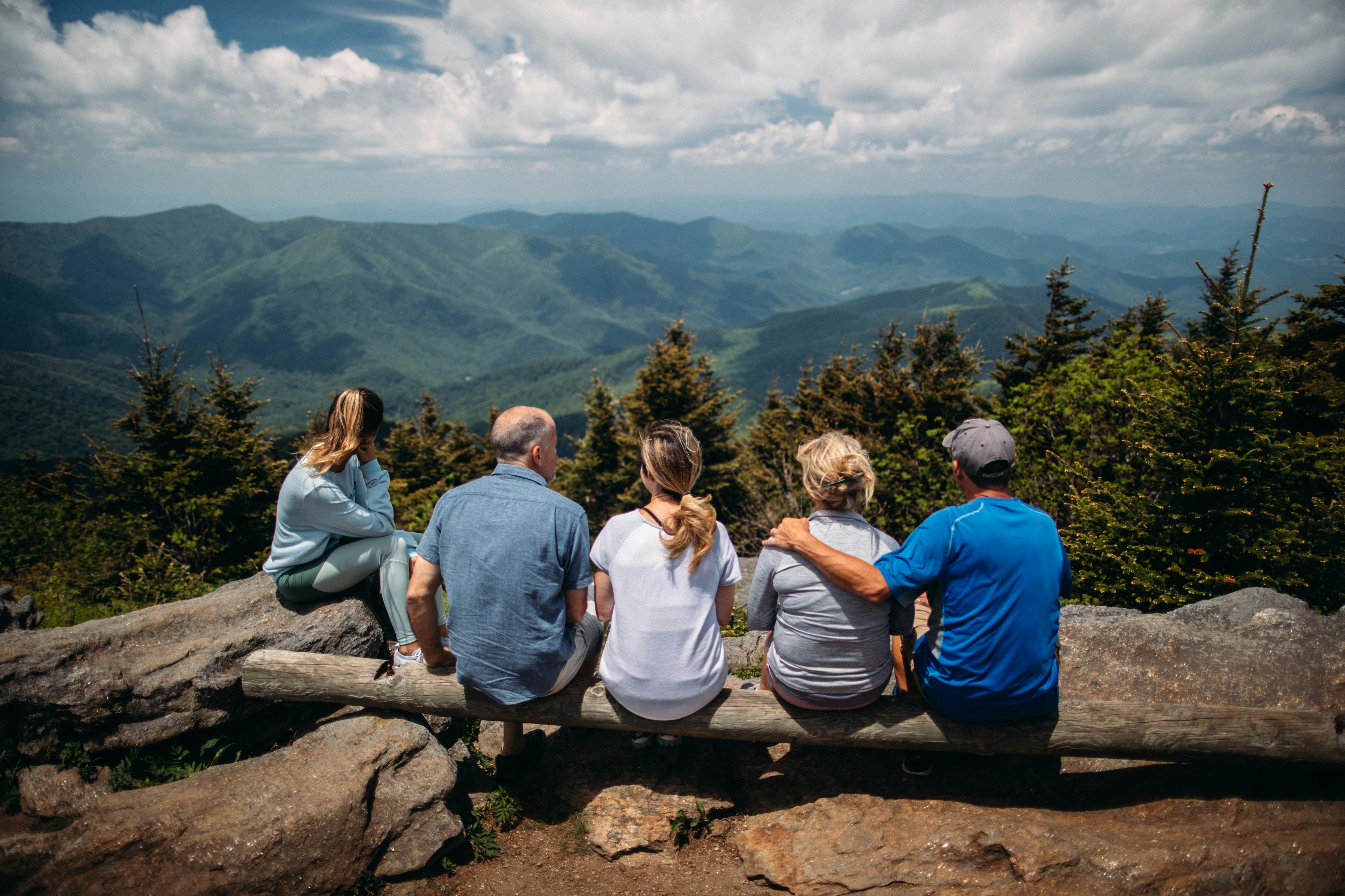 Fünf Personen sitzen nebeneinander auf einem Baumstamm und schauen von einem Berggipfel über eine weite, grüne Berglandschaft