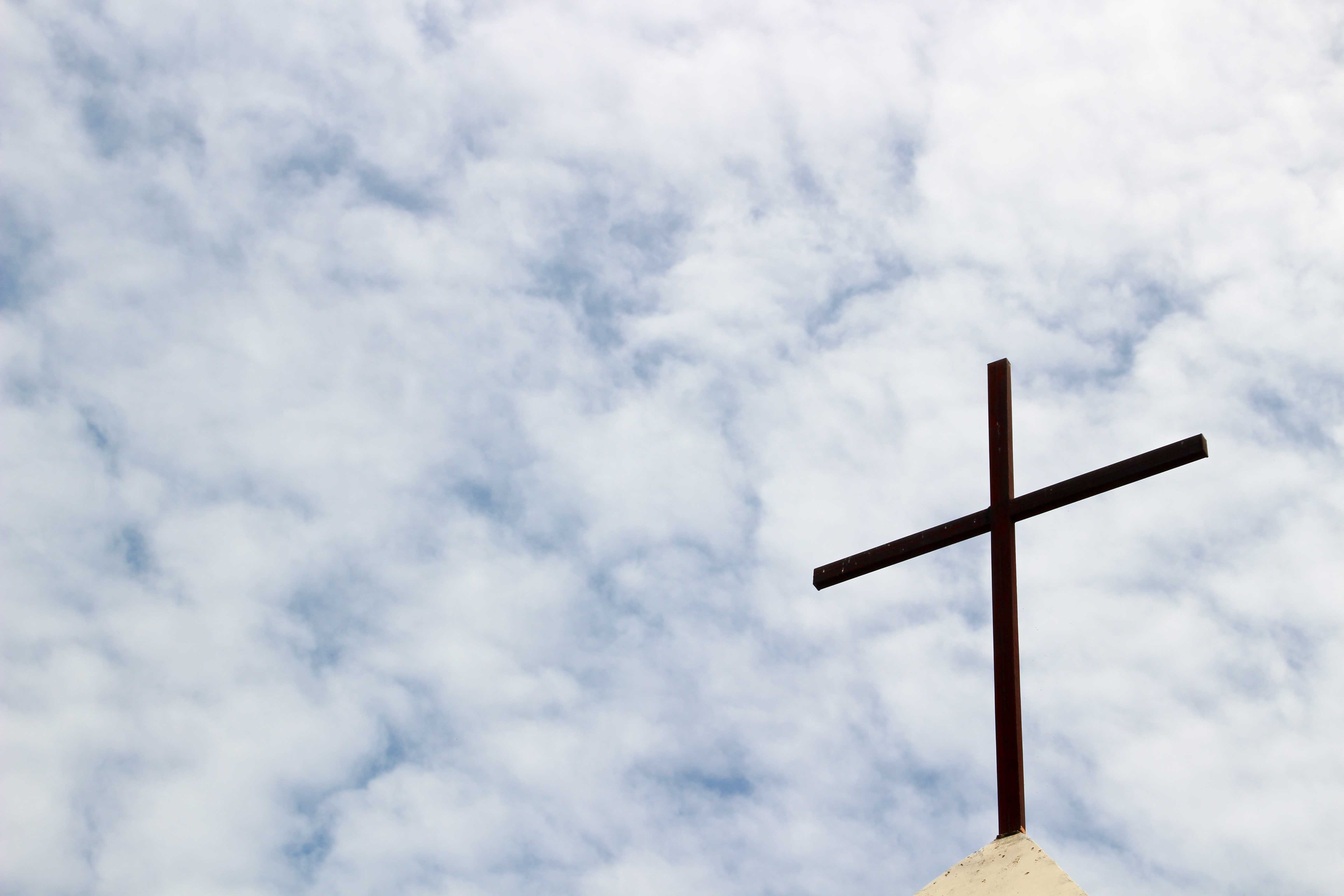 Auf diesem Bild sieht man ein Holzkreuz auf einer Dachspitze. Im Hintergrund ist ein leicht bewölkter Himmel zu sehen.