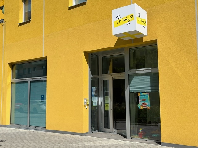 Leuchtend gelbe Hausfassade mit Glaseingang und einem würfelförmigen weißen Schild, auf dem handgeschrieben „Franz“ mit gelben Farbstreifen steht.
