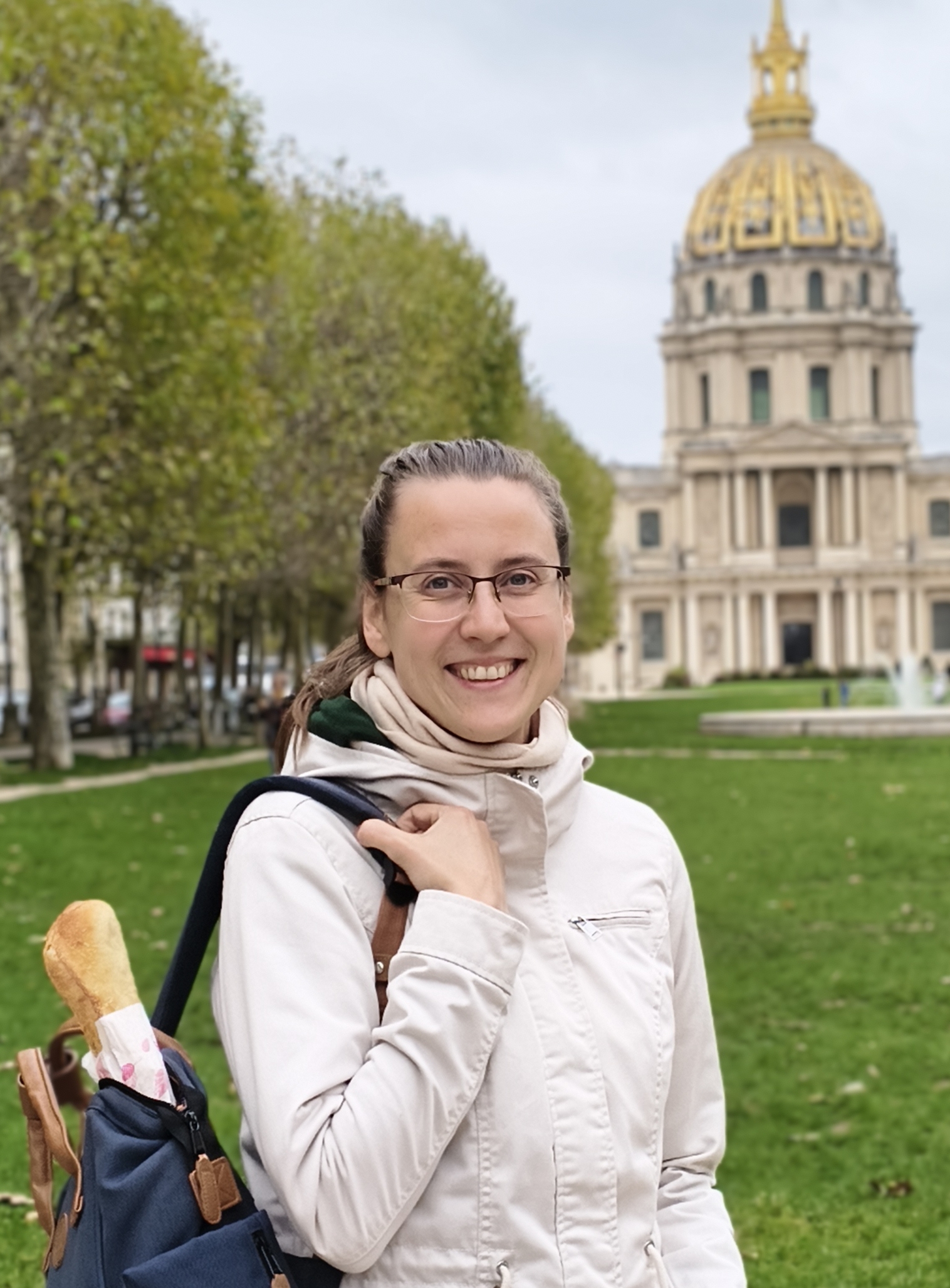 Auf dem Bild sieht man Yasmin Kainer. Sie trägt eine Brille, eine weiße Jacke und aus ihrem Rucksack schaut ein Baguette raus.  Im Hintergrund sieht man eine Kirche und Bäume.