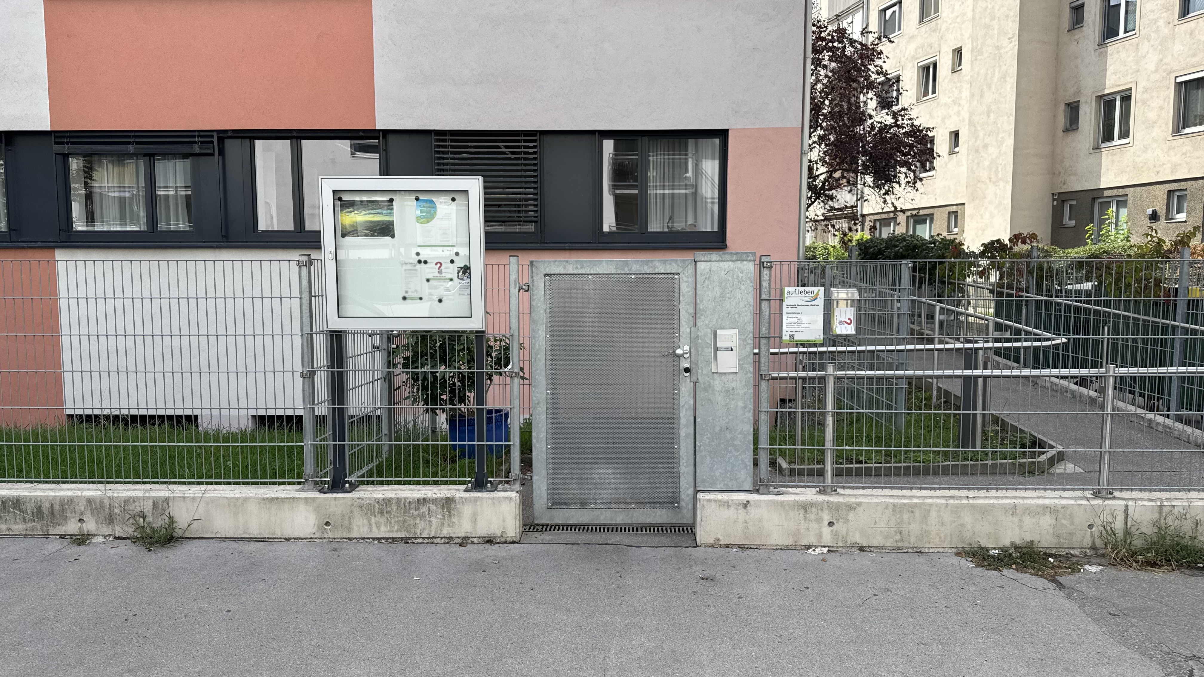 Eingang zur Beratungsstelle: ein verzinktes Metalltor mit Gegensprechanlage, links ein Schaukasten, rechts ein Schild mit der Aufschrift „auf.leben“, davor der Gehsteig