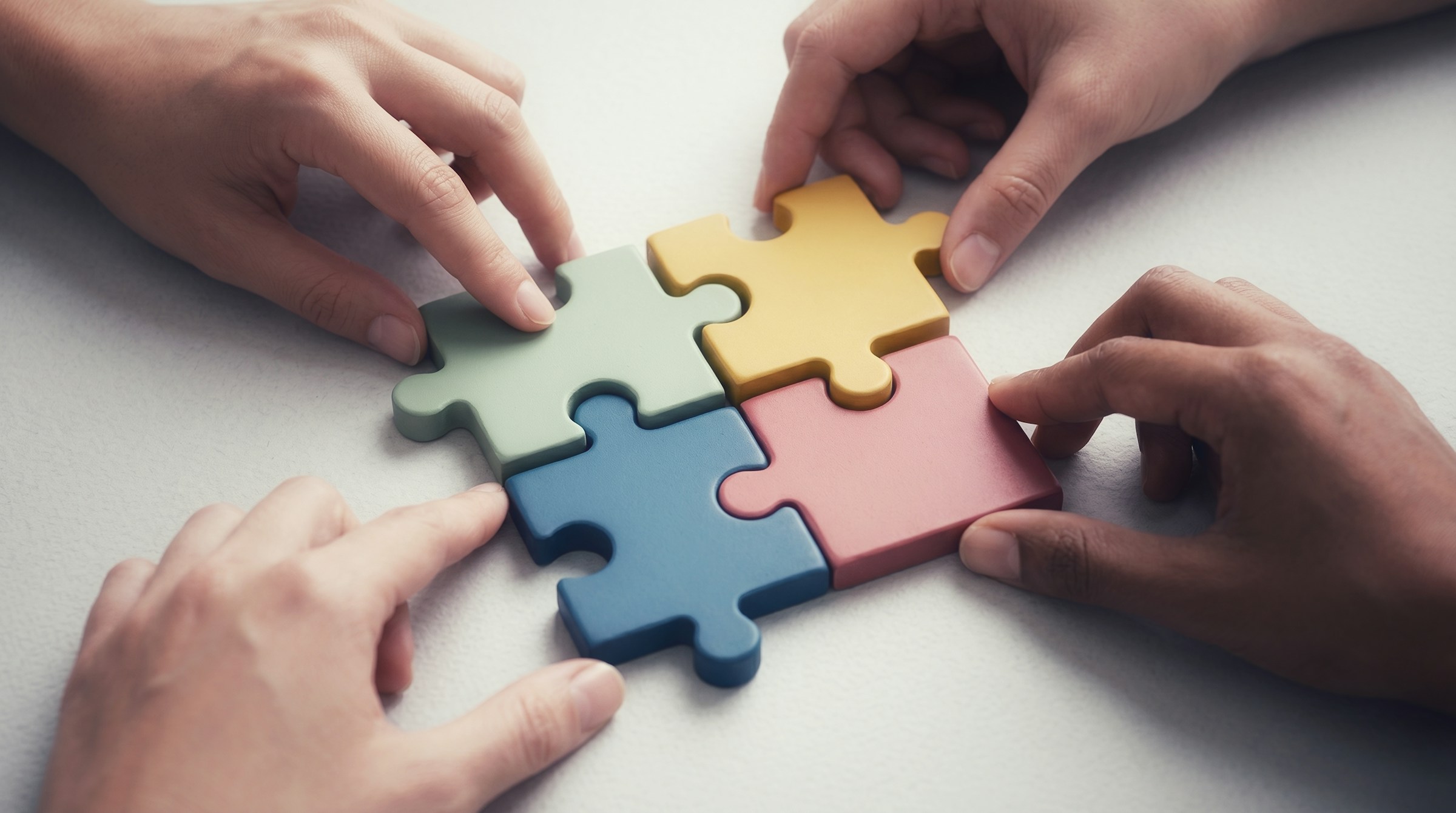 Vier Hände setzen gemeinsam vier bunte Puzzleteile in Grün, Gelb, Blau und Rosa zusammen.