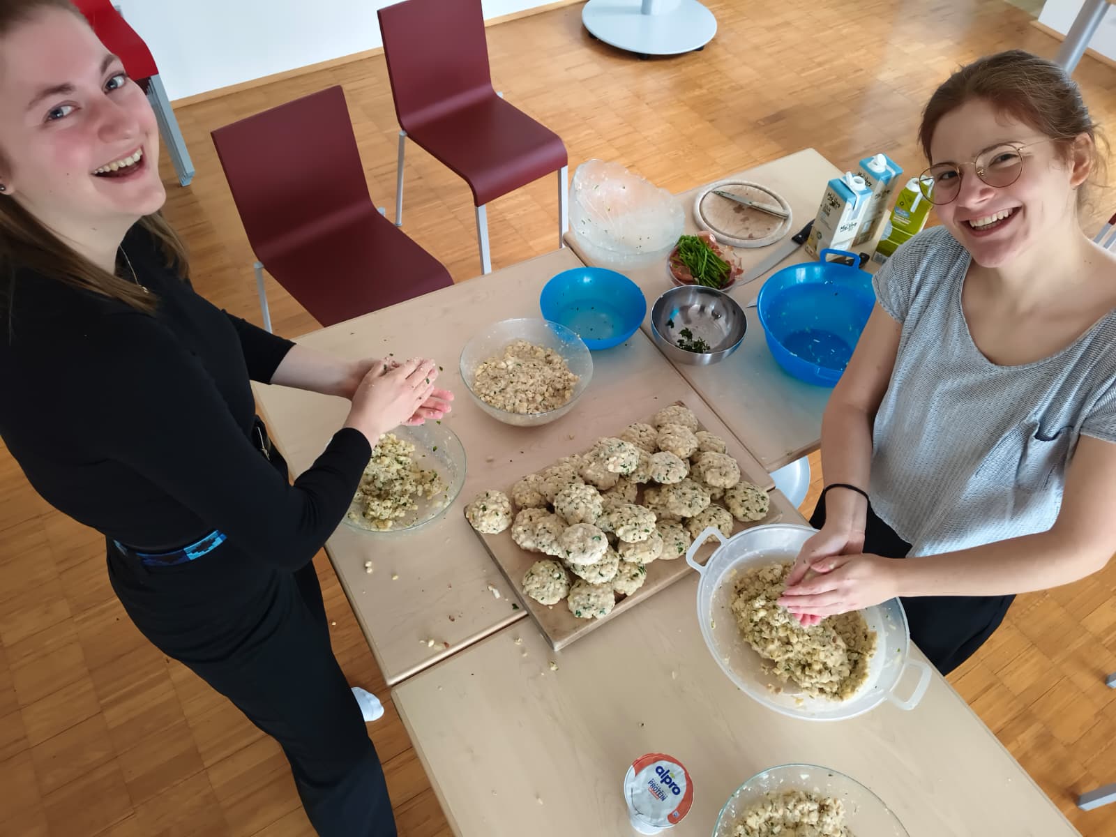 Auf diesem Bild sieht man zwei junge Frauen, die in die Kamera schauen und lächeln. Sie formen gerade auf dem Tisch vor ihnen Knödel, welche sie dann auf eine Platte zwischen sich auf den Tisch ablegen. Auf der Platte liegen schon viele weitere Knöde