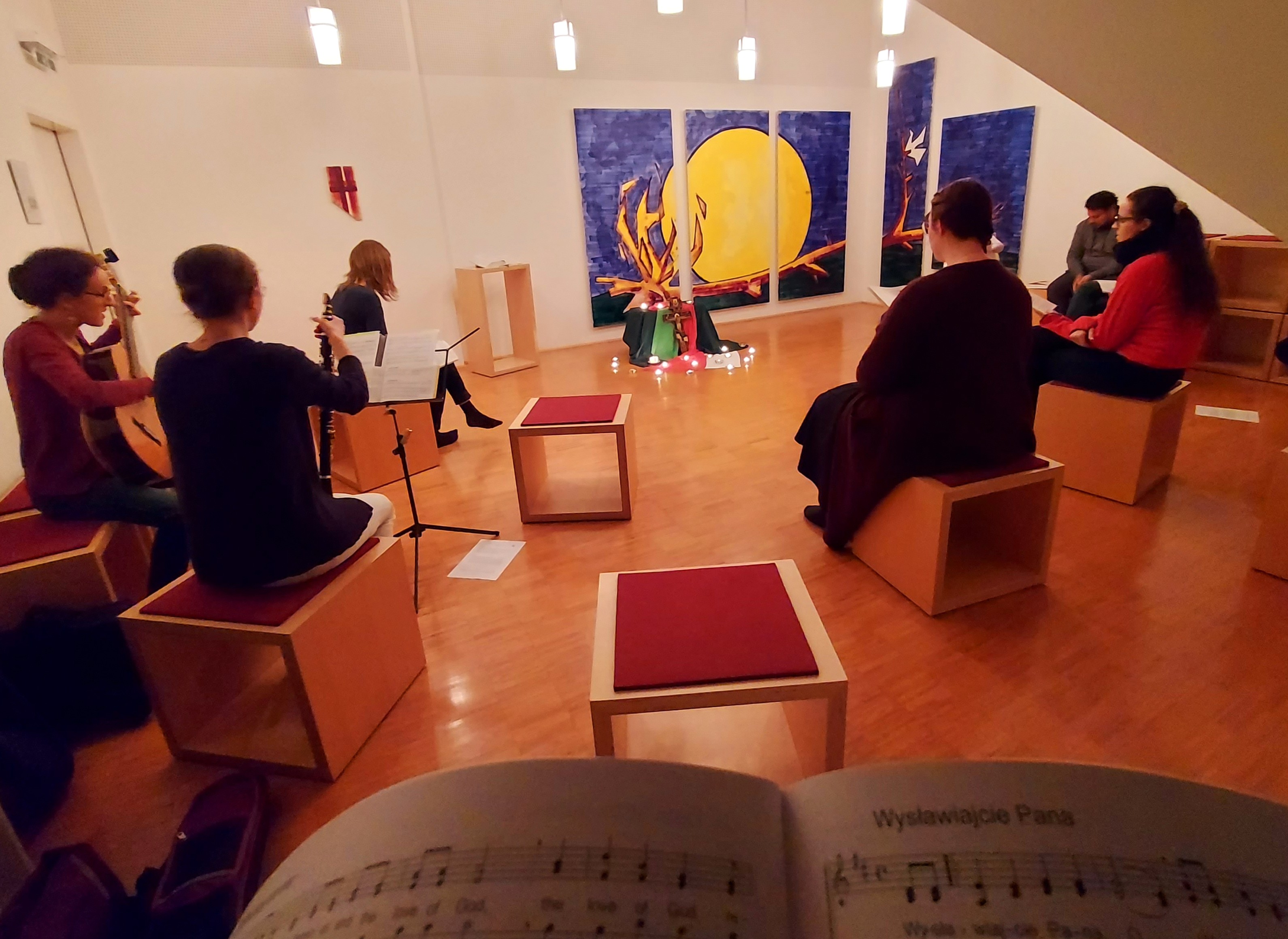 Auf dem Bild sieht man den Andachtsraum vom Zentrum. Im Hintergrund ist an der Wand ein großes Wandbild befestigt und vor diesem stehen Hocker. Auf diesen Hocker sitzen junge Menschen und beten. Eine junge Frau sitzt auf der linken Seite und spielt G
