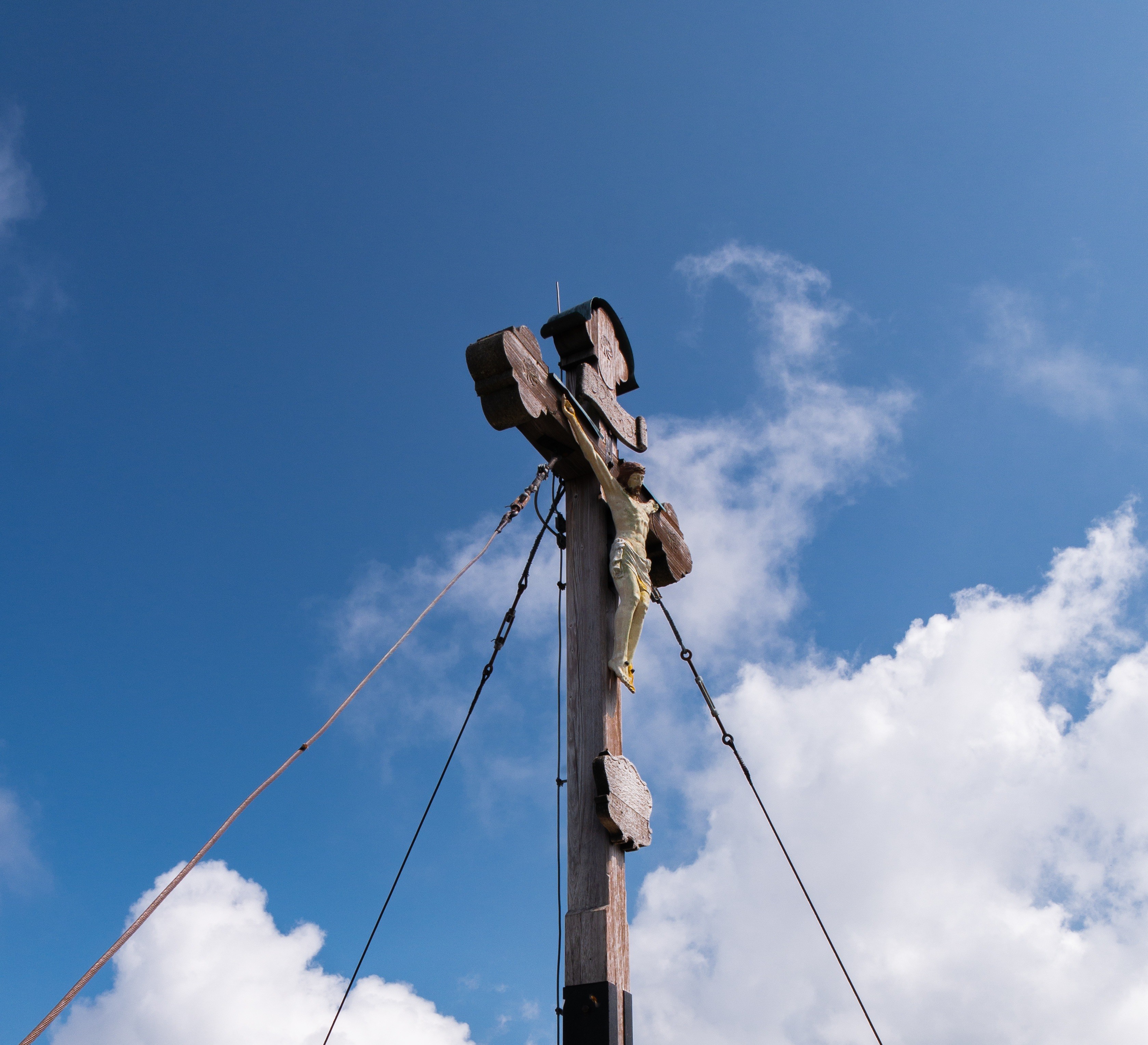 Auf diesem Bild sieht man ein Gipfelkreuz, welches von schräg unten fotografiert wurde. Auf dem kreuz ist Jesus befestigt und das Kreuz wird durch drei Stahlseile gesichert. Im Hintergrund sieht man den blauen Himmel mit paar Wolken.