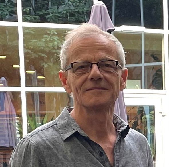 Auf diesem Bild sieht man Thomas Pirkner-Ertl. Er ist ein Mann mit grauen Haaren, einer Brille und ein graues Hemd. Im Hintergrund sieht man eine Fensterscheibe.