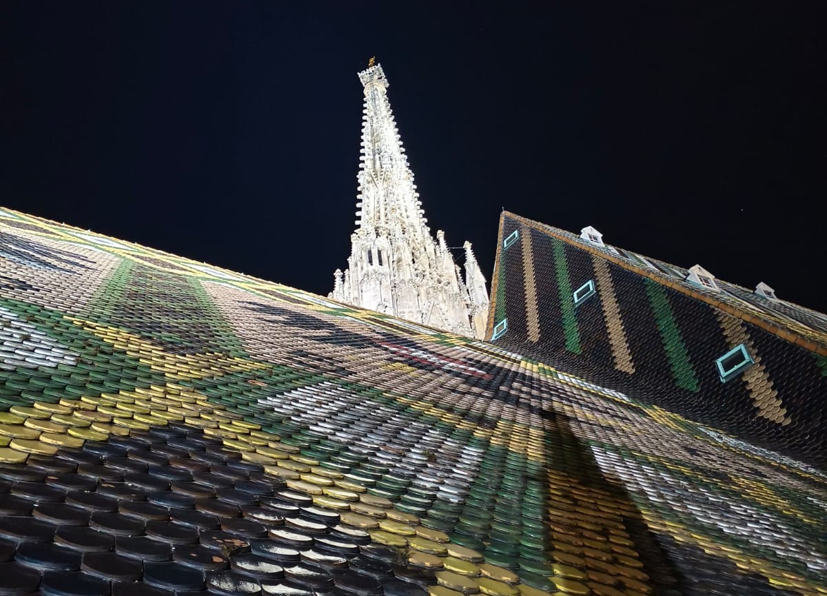 Auf dem Bild sieht man einen Ausschnitt vom Stephansdom bei Nacht. Man sieht die Dachziegel und die Spitze eines Kirchenturm. Der Kirchenturm ist hell beleuchtet.
