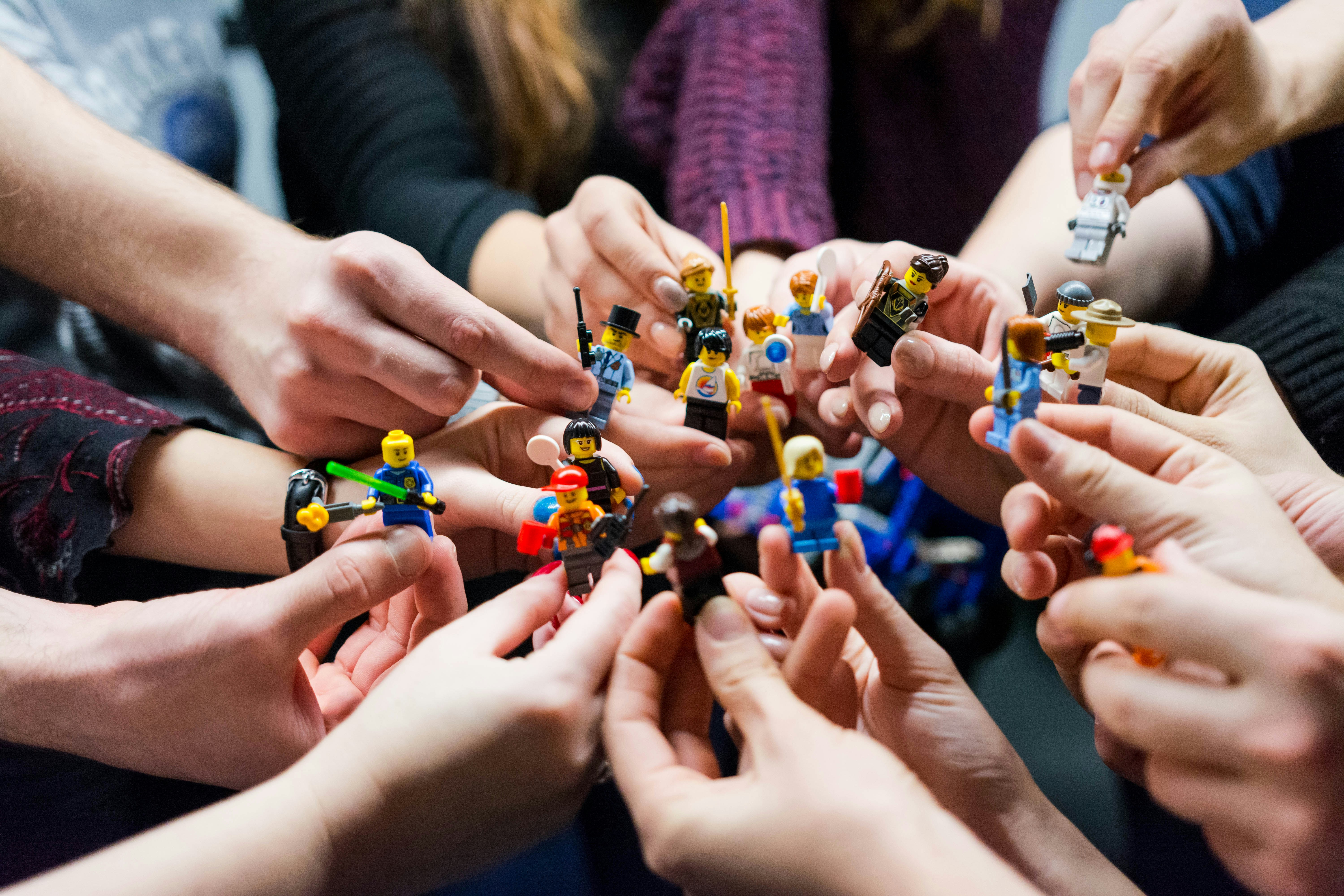 Viele Hände halten in einem Kreis bunte Minifiguren zusammen, jede Figur mit einem anderen Beruf oder Zubehör.