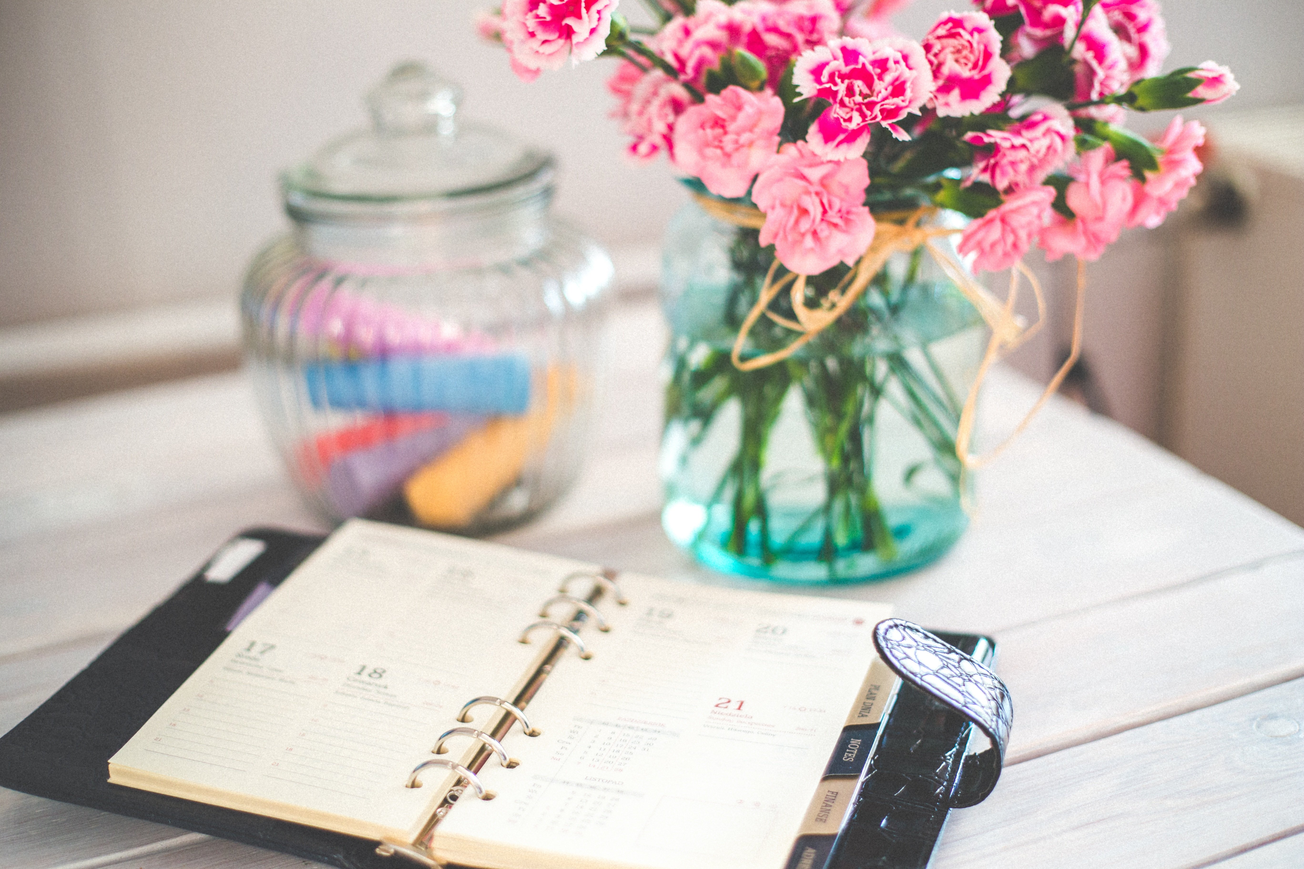 Auf dem Bild sieht man einen offenen Kalender auf einem weißen Tisch liegen und dahinter in einem Glas Textmarker in verschiedenen Farben und einen Strauß rosa Blumen in einer Vase.