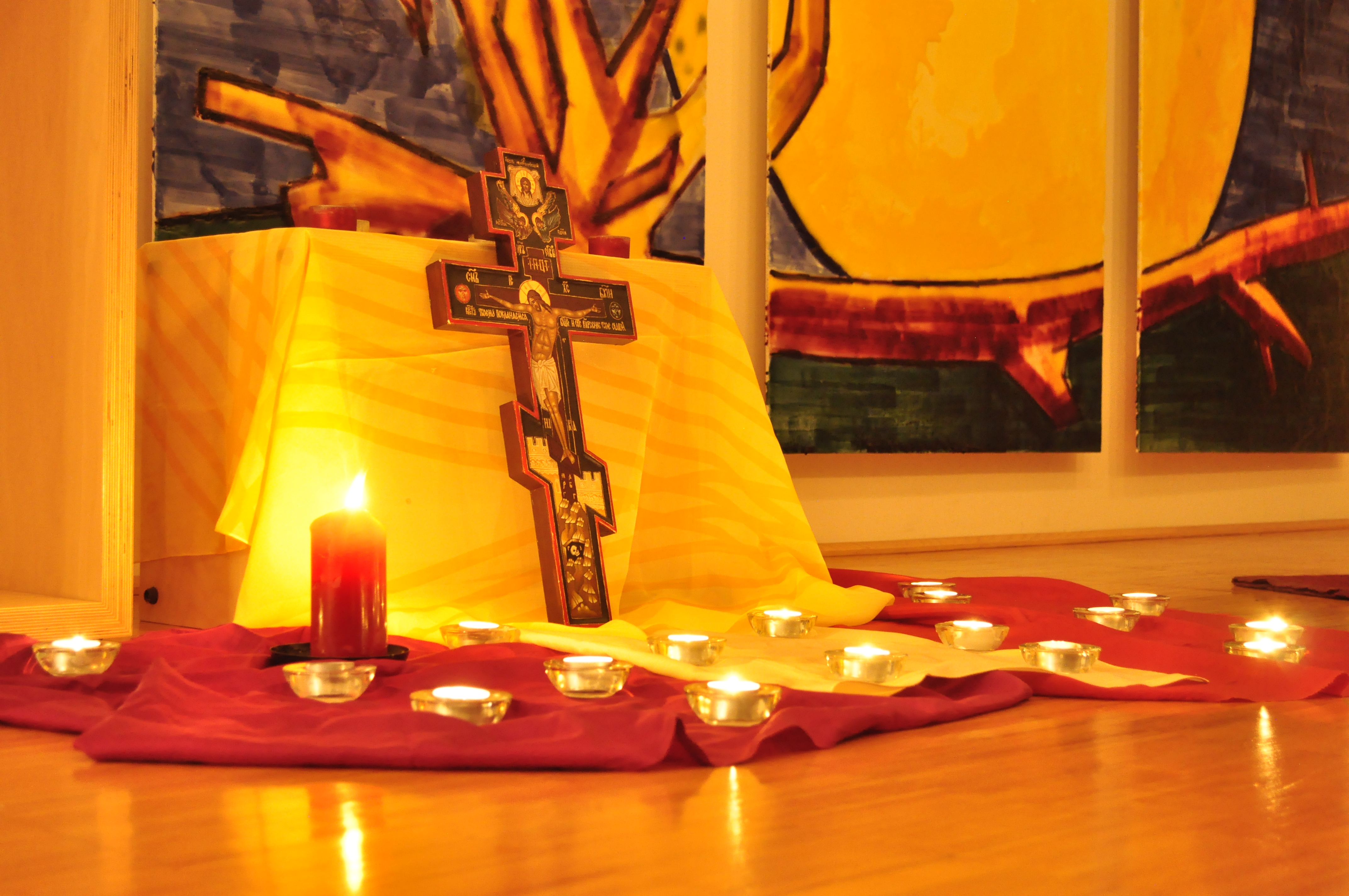 Auf dem Bild sieht man ein Kreuz an einen kleinen Tisch mit einer Tischdecke drüber, angelehnt. Vor dem Kreuz ist ein rotes Tuch ausgebreitet und brennende teelichter stehen drauf. Im Hintergrund sieht man ein Ausschnitt von dem Wandbild, was im Anda
