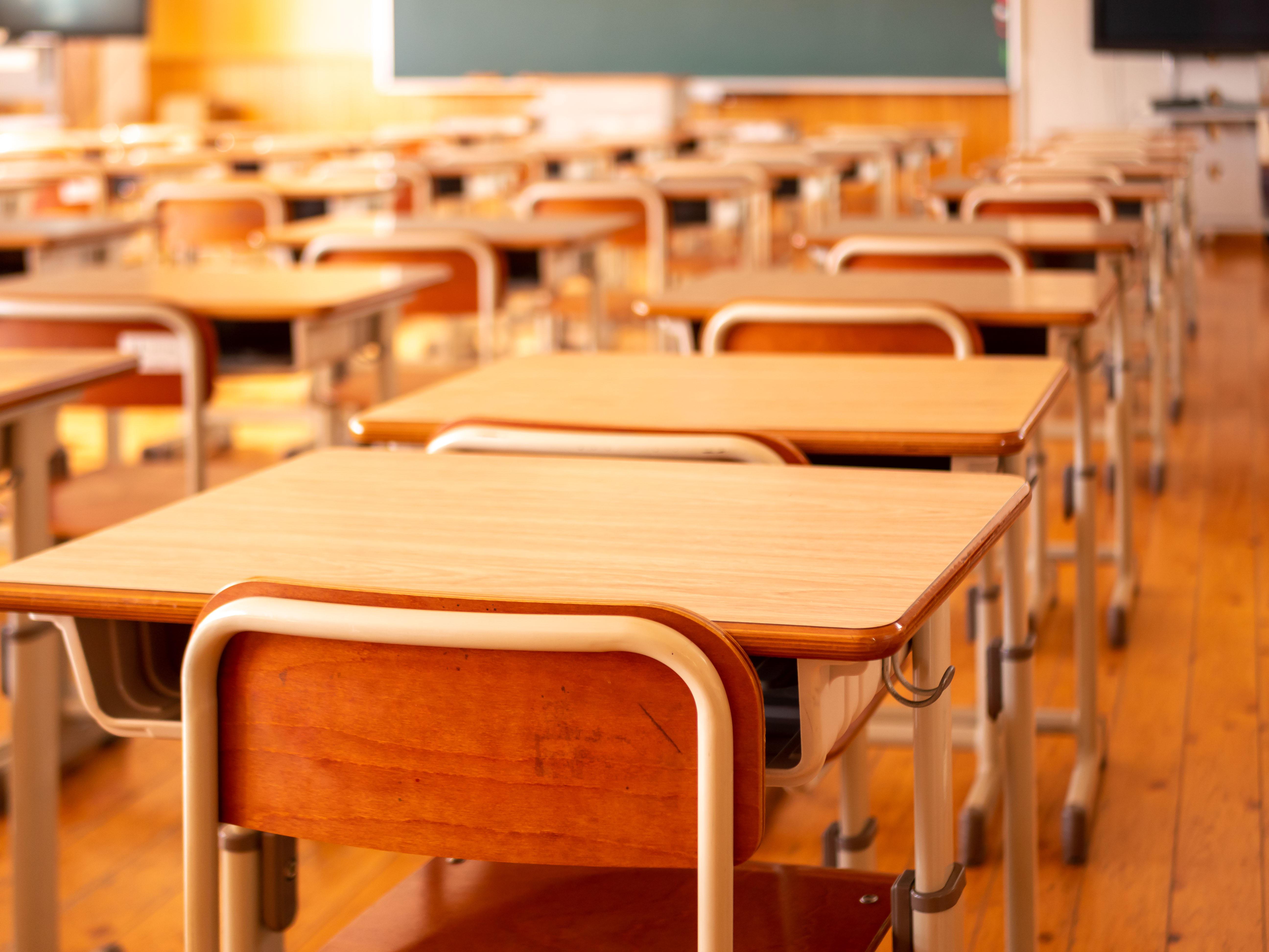 In diesem Bild sieht man ein leeres Klassenzimmer. Es sind SHolzstühle und Tische vor einer Tafel abgebildet.