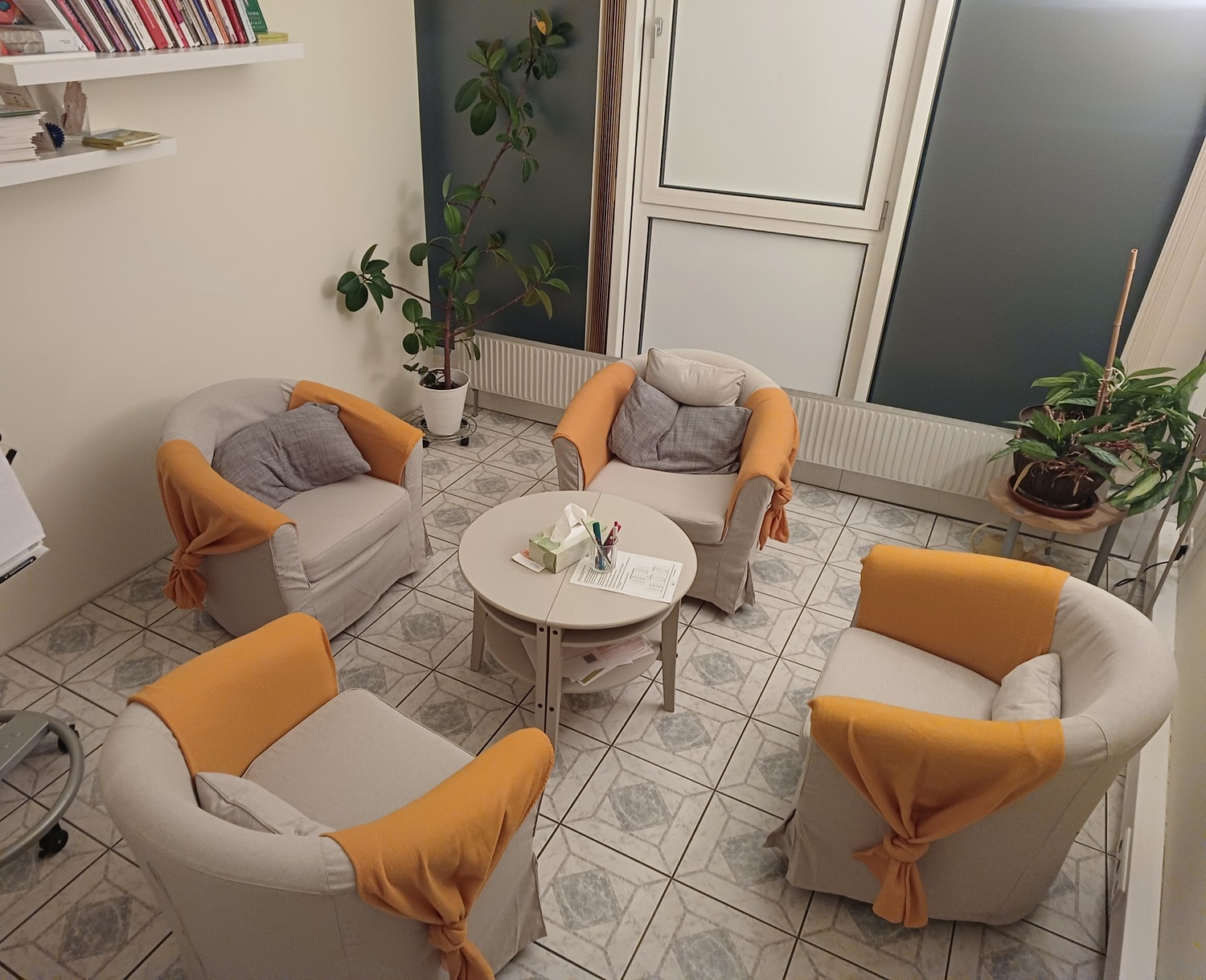 Heller Gesprächsraum mit vier beigen Sesseln mit orangefarbenen Decken um einen runden Tisch, dazu Pflanzen und ein Bücherregal