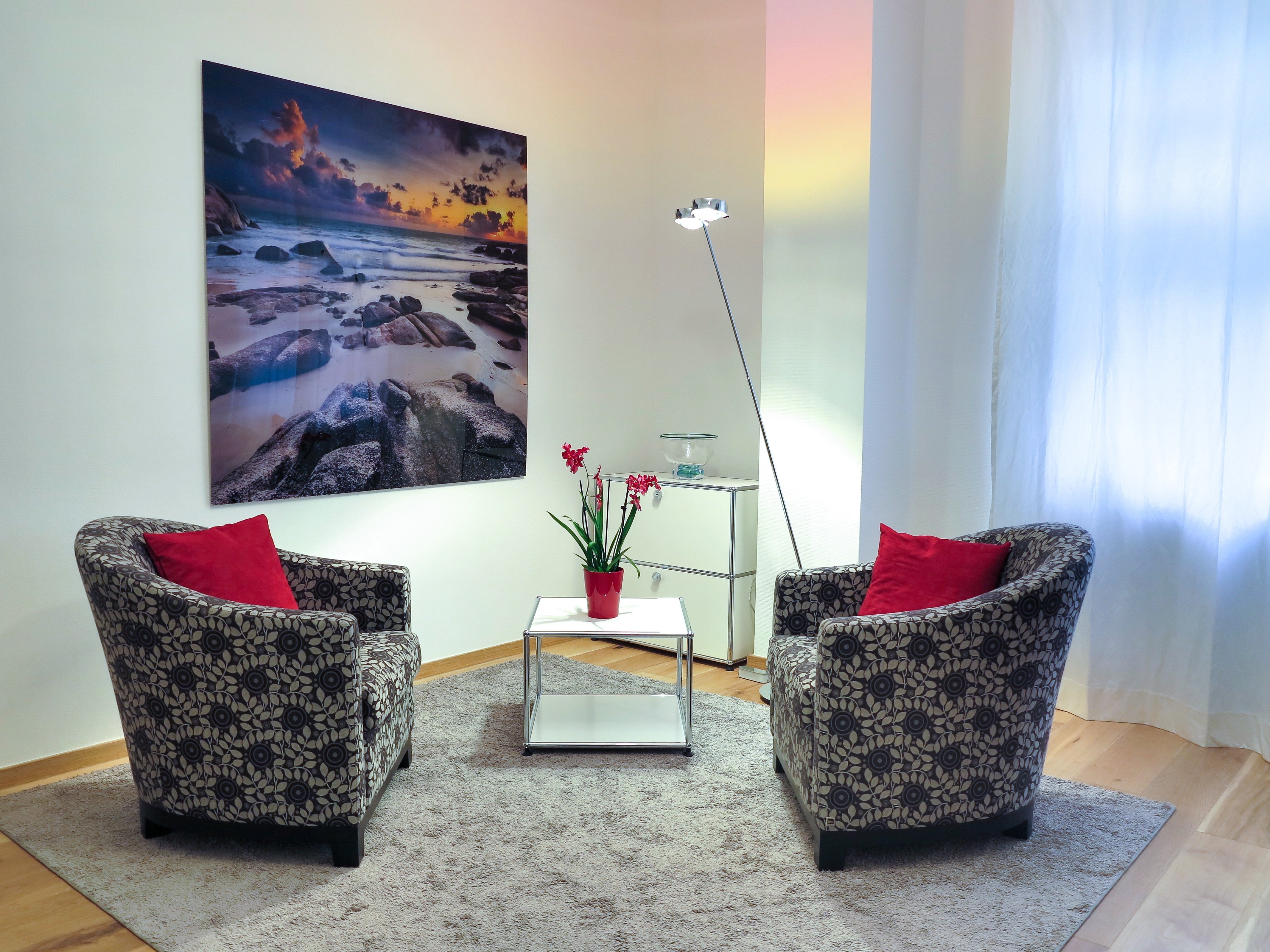 Gemütlich eingerichteter Beratungsraum mit zwei gemusterten Sesseln mit roten Kissen, einem kleinen Beistelltisch mit Orchidee und einem großformatigen Strandfoto an der Wand