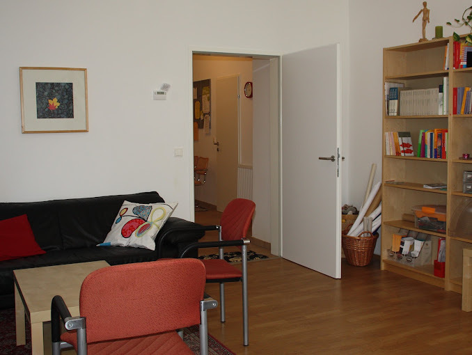 Heller Raum mit schwarzem Sofa, mehreren Stuehlen, kleinem Tisch, offenstehender Tuer und Buecherregal.