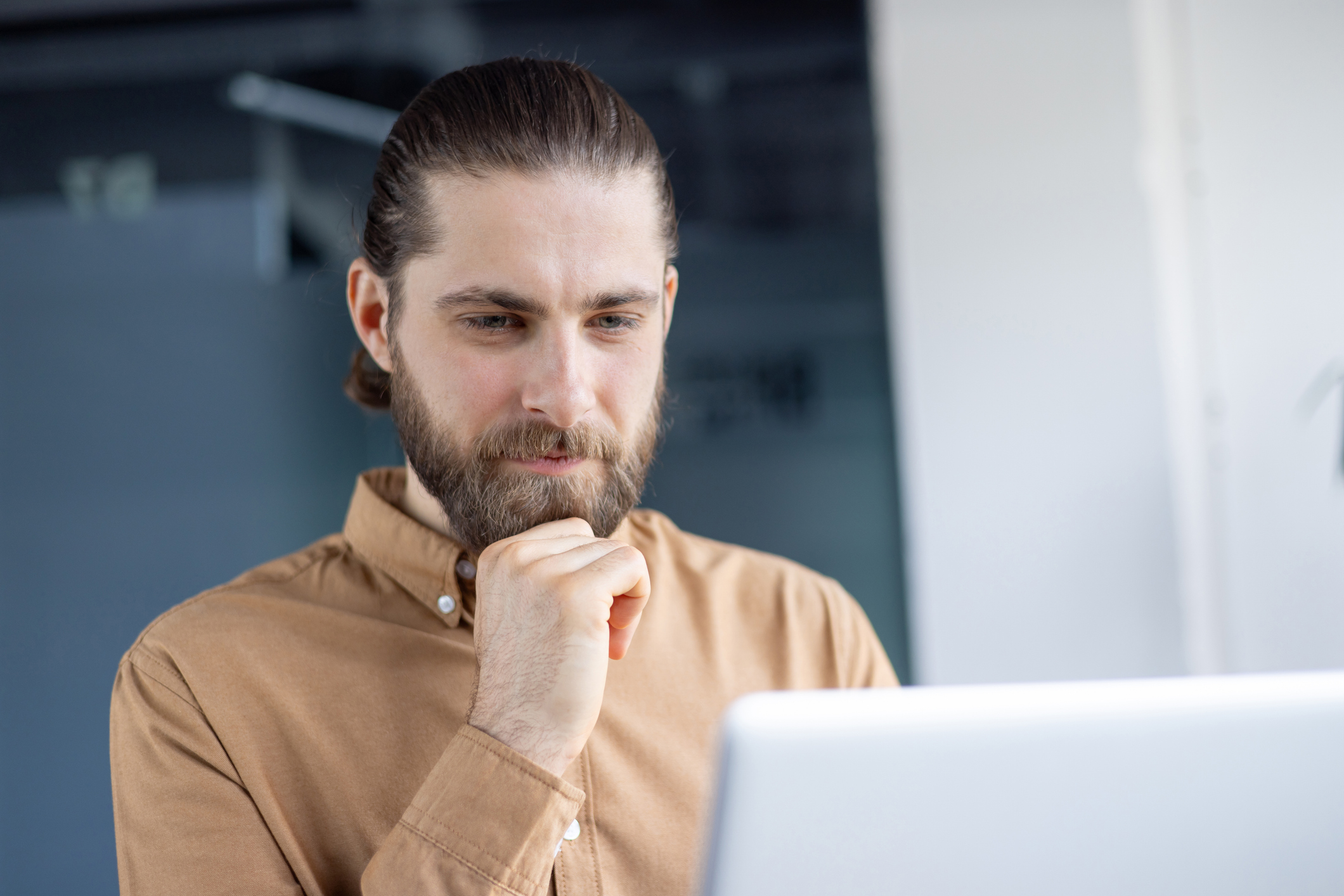 A bearded man with a ponytail looks down at a laptop, pondering a thought while working in an office setting.