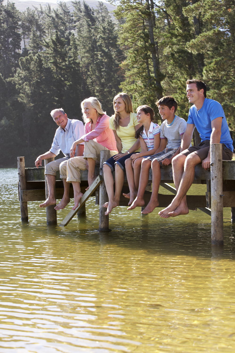 Smiling And Laughing Three Generation Family Sitting On Wooden Jetty Looking Out Over Lake.