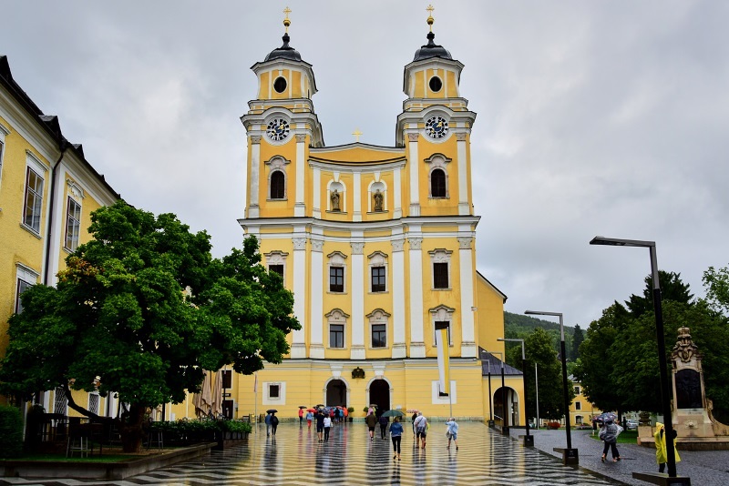 Mondsee, Austria - 05.08.2016: Basilica St. Michael, a yellow and white church building, a clock is installed on the towers, the square is covered with white and black tiles, trees grow nearby.