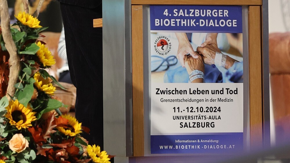 Aufgehängtes Veranstaltungsplakat zu den Salzburger Bioethik-Dialogen 2024 gereichten Händen als Bild
