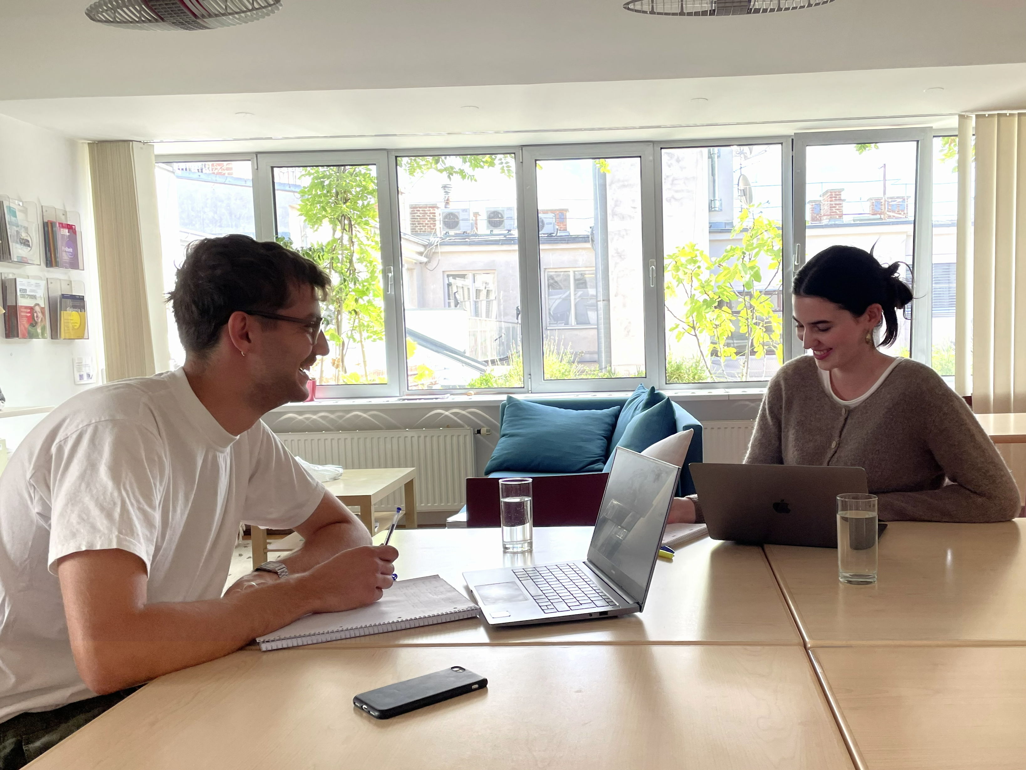 Auf diesem Bild sieht man zwei junge Menschen, einen Mann und eine Frau, wie sie an einem Tisch lernen. Der Mann sitzt vor seinem Laptop und schreibt etwas in sein Notizbuch. Die Frau hat ein Tablet in der Hand und schaut auf diese. Im Hintergrund si