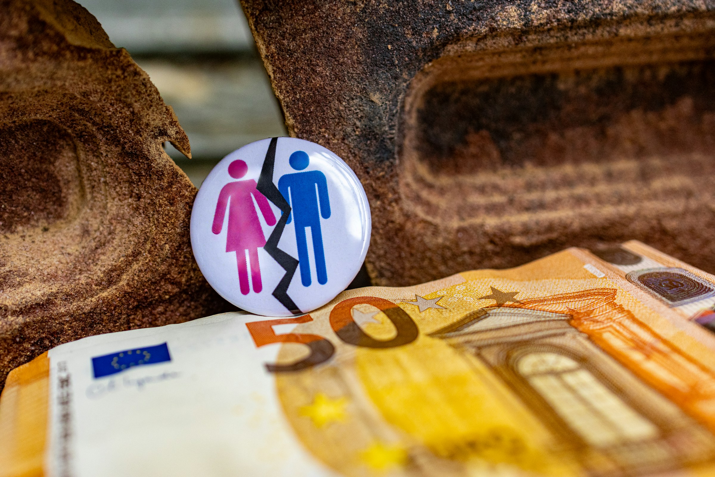Ein Anstecker mit einer pinken Frauen- und einer blauen Männerfigur, getrennt durch einen schwarzen Zickzack-Riss, lehnt zwischen Sandsteinfelsen an einem 50-Euro-Schein