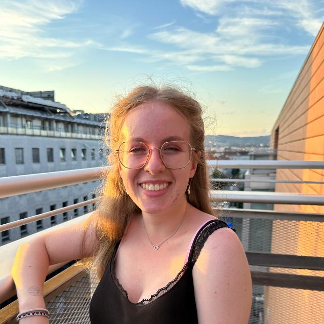 Auf dem Bild sieht man Hannah Greiner, eine Frau mit blonden Haaren, einer Brille und einem schwarzen Trägertop. Im Hintergrund sieht man Häuser und den blauen Himmel  und Hannah Greiner steht auf einem Balkon.